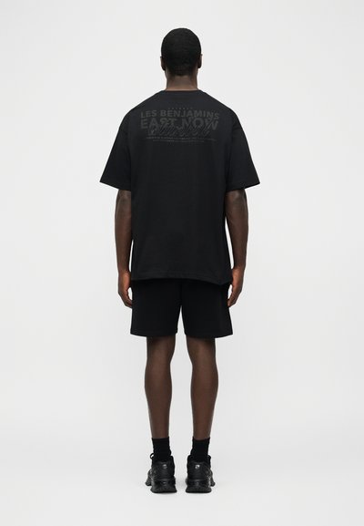 Άνδρας που στέκεται με πλάτη, φοράει μαύρο φαρδύ μπλουζάκι με μικρό κείμενο τυπωμένο και μαύρο σορτς, μπροστά σε απλό ανοιχτόχρωμο φόντο.