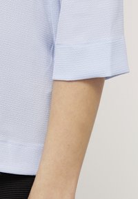 Camicetta azzurro chiaro con texture e maniche arrotolate, che mostrano una superficie di tessuto liscia e un motivo sottile. Nessun hardware o dettagli visibili.