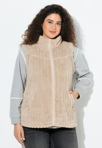 Gilet en polaire beige avec col montant et fermeture éclair à l'avant. Il présente une texture côtelée et des poches. Porté par-dessus une chemise à manches longues grise.