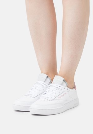 Nogi w białych niskich butach Reebok z różowymi akcentami, stojące na jasnym, jednolitym tle.