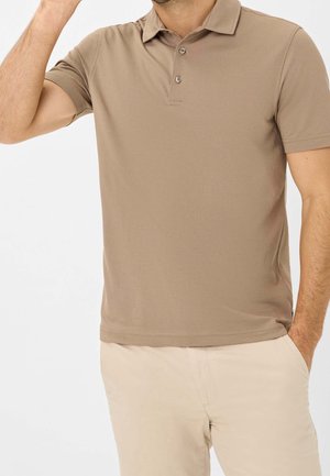 Homme portant un polo beige à manches courtes et un pantalon beige clair, une main dans la poche, l'autre levée près de la tête.