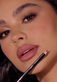 Kredka do ust z naostrzonym końcem, trzymana w pobliżu pełnych, matowych, cielistych warg. Delikatny, neutralny makijaż oczu oraz wyraźnie podkreślone brwi uzupełniają wygląd.