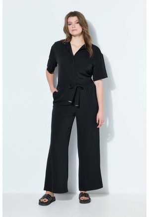 Junge Frau mit langen Haaren steht vor einer weißen Wand, trägt einen schwarzen Overall mit kurzen Ärmeln und weitem Bein sowie schwarze Sandalen.