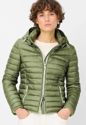 Woman with short curly hair wearing an olive green quilted hooded jacket, hands in pockets, against a plain light background.