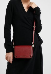 Persona in un vestito nero che tiene una borsa a tracolla in pelle rossa con hardware dorato e tracolla regolabile sulla spalla.