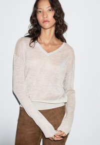 Woman with curly dark hair wearing a light beige V-neck sweater and brown pants, standing against a plain white background.