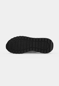 Semelle en caoutchouc noire d’une chaussure avec des crampons rectangulaires et des rainures, montrant le motif de la semelle inférieure et le nom de la marque "Guess" au centre.