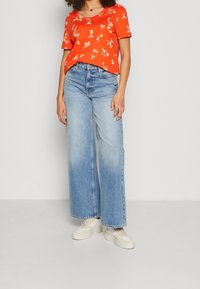 Rød blomstermønstret kortærmet top, parret med lyseblå vidde jeans og cremede sneakers. Har en afslappet pasform og V-hals design.