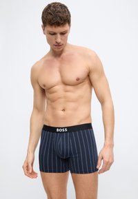 Μυώδης άνδρας χωρίς πουκάμισο, φοράει σκούρα μπλε boxer με ρίγες, μαύρη ζώνη μέσης και λευκό λογότυπο "BOSS", στέκεται μπροστά σε απλό φόντο.