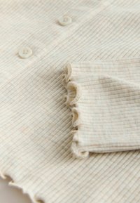 Tissu tricot côtelé crème avec petits boutons et bords froncés sur la manche et l'ourlet d'un vêtement.