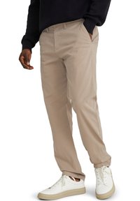 Homme portant un pantalon beige, des baskets blanches avec des semelles beiges, et un pull noir, debout avec une main dans la poche.