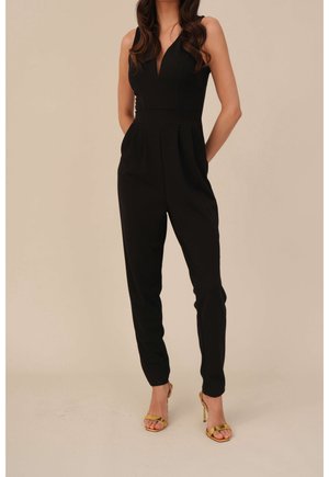 Vrouw in een mouwloze zwarte jumpsuit met een diepe V-hals en geplooide broekspijpen, gecombineerd met gouden hoge hakken op een beige achtergrond.