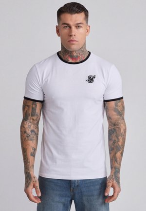 Mann mit Tattoos am Hals und an den Armen trägt ein weißes T-Shirt mit schwarzem Rand und kleinem Logo auf der Brust, steht vor einem einfarbigen hellen Hintergrund.