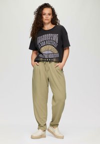 Zwarte graphic t-shirt met tekst en zonontwerp; olijfgroene broeken met een relaxte pasvorm en elastische tailleband; lichtgekleurde instap schoenen.