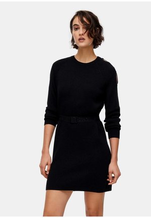 Donna che indossa un vestito maglione nero a maniche lunghe con cintura e un piccolo dettaglio a taglio sulla spalla sinistra, in piedi davanti a uno sfondo semplice.