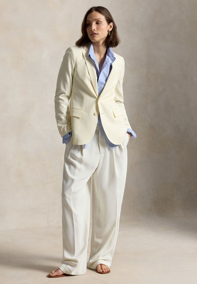 Nő krémszínű blézert és széles szárú krémszínű nadrágot visel világoskék inggel, zsebre tett kézzel áll egyszínű háttér előtt.