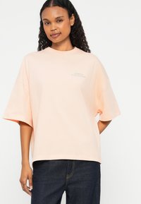 T-shirt oversized de cor pêssego, feito de um tecido suave, com mangas curtas e um logótipo simples impresso numa cor contrastante no peito.