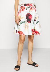 Vit blommig kjol med röda, rosa och gula blommor, utsvängd design, ett åtsittande midjeband och slät tygtextur. Svarta sandaler bärs.