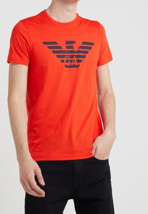 Rotes Baumwoll-T-Shirt mit einem auffälligen navyblauen Logo mit Flügeln auf der Vorderseite. Kurze Ärmel und klassischer Rundhalsausschnitt.