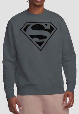 Mann trägt ein dunkelgraues Sweatshirt mit einem großen schwarzen Superman-Logo auf der Brust, kombiniert mit hellbeigen Hosen.