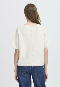 Witte lichtgewicht gebreide trui met korte mouwen en een ronde halslijn. Voorzien van een knoopsluiting aan de achterkant. Gepaard met denim jeans.