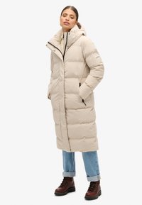 Giacca a vento beige con cappuccio, dotata di chiusura con cerniera, tasche laterali e un design trapuntato. Abbinata a jeans blu arrotolati e stivali marroni.
