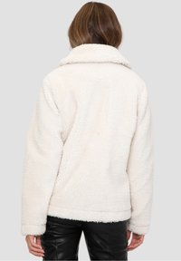 Veste en fausse fourrure couleur crème avec un col haut, surface texturée et coupe décontractée, associée à un pantalon en cuir noir.