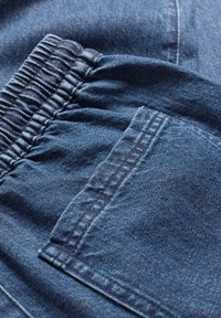 Denim-kangasta tummansinisen sävyisenä, jossa on kerätty vyötärö ja yksi sivutasku. Ompeluyksityiskohtia näkyvissä reunoilla.