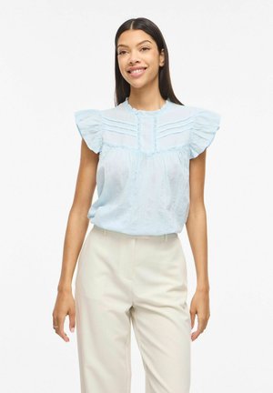 Lächelnde Frau mit langen dunklen Haaren, die eine hellblaue, mit Rüschen verzierte Bluse und cremefarbene Hosen mit hoher Taille trägt, steht vor weißem Hintergrund.