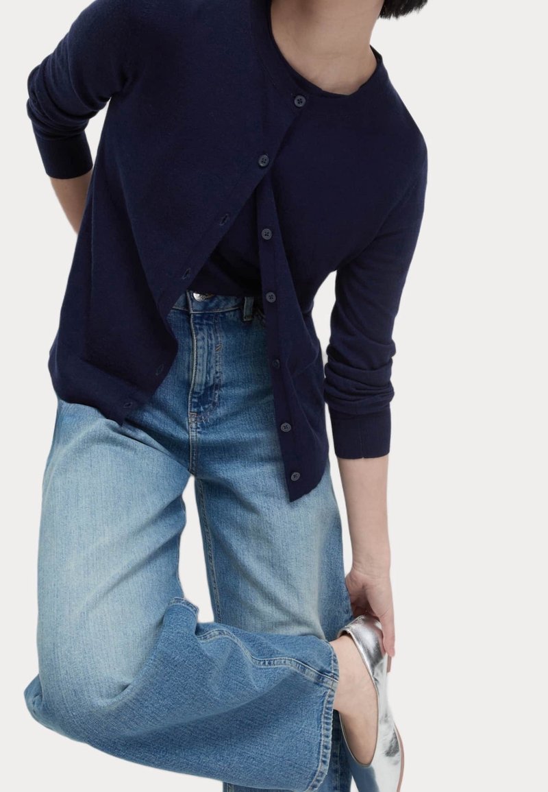 Marinefarbener Cardigan mit Knöpfen, über einem schwarzen Oberteil getragen, kombiniert mit hellblauen weit geschnittenen Jeans und silbernen Schuhen. Glatte Textur und lässiger Stil.