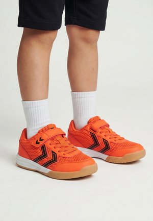 Kind draagt feloranje sportschoenen met zwarte chevronpatronen, witte sokken en zwarte korte broek op een lichte achtergrond.