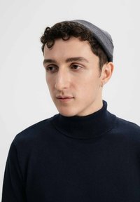 Navy coltrui van zacht materiaal, met een geribde kraag. Een grijze gebreide beanie rust op kort, krullend haar.