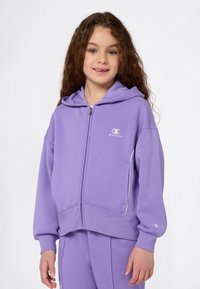 Ragazzina con lunghi riccioli che indossa una felpa con zip viola della Champion e pantaloni abbinati, sorridente contro uno sfondo bianco.
