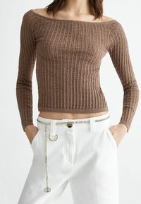 Nainen, joka on pukeutunut ruskeaan ribbikuvioiseen olkaimeton pitkähihaiseen puseroon ja valkoisiin korkeavyötäröisiin housuihin, joissa on ohut hopeaketju vyönä, kädet taskuissa.