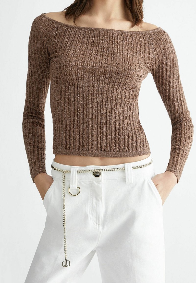 Nainen, joka on pukeutunut ruskeaan ribbikuvioiseen olkaimeton pitkähihaiseen puseroon ja valkoisiin korkeavyötäröisiin housuihin, joissa on ohut hopeaketju vyönä, kädet taskuissa.