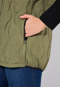 Gilet verde oliva trapuntato con motivo a onde testurizzate, dotato di una tasca laterale con zip e orlo elasticizzato. Indossato sopra una maglietta nera a maniche lunghe.