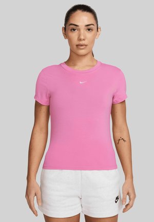 Donna con i capelli scuri legati indietro, che indossa una maglietta rosa Nike e pantaloni corti bianchi Nike, in piedi contro uno sfondo grigio semplice.