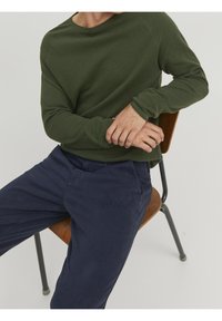 Grön waffle-stickad långärmad tröja i kombination med marinblå bomullsbyxor. Tröjan har en rund halsringning och åtsittande ärmslut. Bakgrunden består av en trästol.