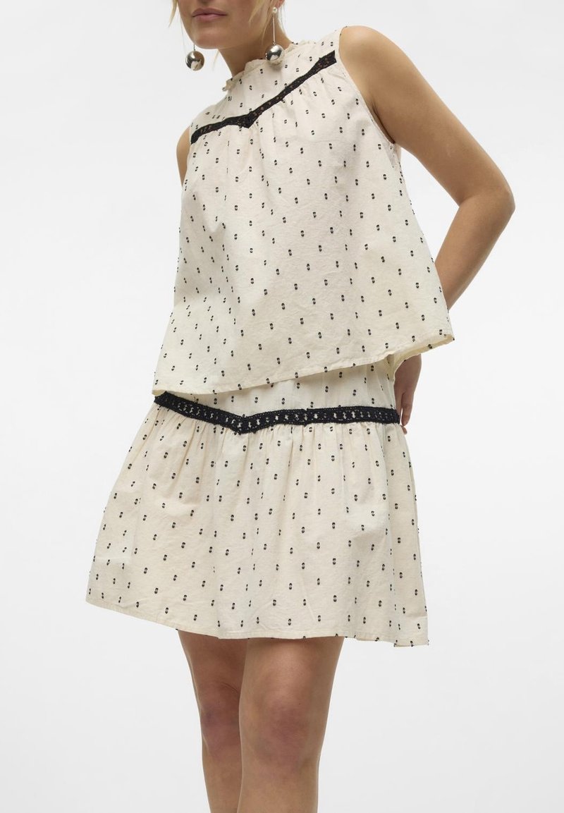Femme portant une robe crème sans manches avec de petits pois noirs et une bordure en dentelle noire, debout sur un fond blanc uni.