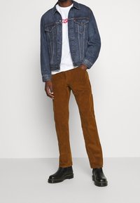 Chaqueta de mezclilla, camiseta blanca con logo rojo, pantalones de pana marrón y botines negros. Las características incluyen un corte clásico y un tejido texturizado.