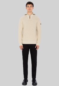 Beiger Strickpullover mit einem Viertel-Reißverschlusskragen, geripptem Muster und kontrastierendem Innenkragen. Kombiniert mit schwarzen Jeans und Schuhen.