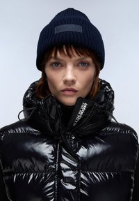 Doudoune noire et brillante avec col haut et fermeture éclair, associée à un bonnet en tricot bleu marine avec un patch logo texturé.
