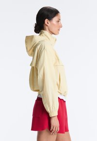 Femme aux cheveux foncés en chignon, portant une veste à capuche jaune clair et un short rouge, debout de profil sur fond blanc.