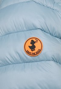 Rund orange plet med en sort syngende and og teksten "RED DANDEN" på lyseblå quiltet stof.