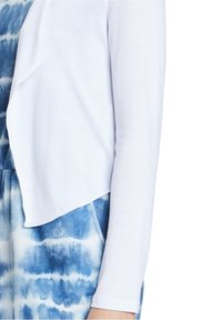 Weißer Strickcardigan mit einem fließenden Schnitt, kombiniert mit einem blau-weißen Batik-Overall mit einem Rundhalsausschnitt und seitlichen Taschen.