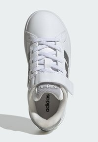 Bílá sportovní bota s hladkým koženým svrškem, zavazováním na tkaničky a suchý zip, polstrovaným límcem a šedou textilní podšívkou; má tři černé pruhy.