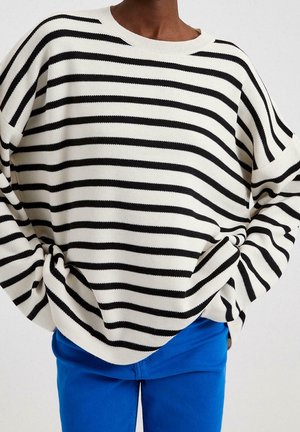 Person iført en løs hvid sweater med sorte vandrette striber og lyseblå bukser, hænderne gemt inde i sweaterens sider.