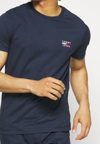 Marineblå bomullströja med rundad halsringning och korta ärmar. Har en vit och röd "TOMMY JEANS" logotyp på övre vänstra sidan. Slät textur.