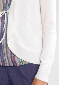 Cárdigan blanco de punto con puños acanalados, que cuenta con un cierre de corbata. Debajo, asoma una blusa estampada en tonos de azul, verde y rosa.