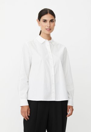 Kvinde med mørkt hår sat op i hale iført en hvid skjorte med knapper og sorte bukser, stående foran en ensfarvet hvid baggrund.